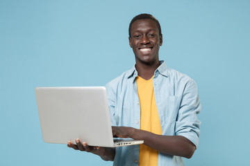 Smiling young african american man guy in casual shirt, yellow t-shirt posing isolated on blue background studio portrait. People lifestyle concept. Mock up copy space. Working on laptop pc computer.