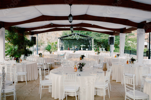 Wedding dinner table reception. Round tables with white tablecloths and white Chiavary chairs under a large white tent, against the backdrop of a musical group.