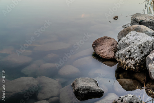 Canvas Print Stones and rocks beneath the very still surface of a misty lake, and the rocky shore of reddish and white rocks offset to the left of the unmoved surface of water