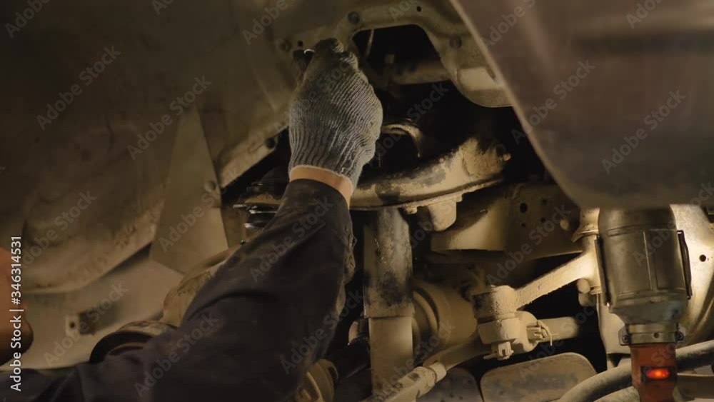 An Asian man is repairing the chassis of an old car lifted on a lift in a workshop, close-up. An Asian man is repairing the chassis of an old car lifted on a lift in a workshop, close-up.