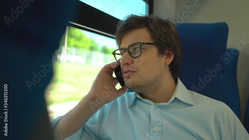 Handsome young businessman wearing a blue shirt and glasses is riding a train dialing a number and talking on his phone. Sunny summer day