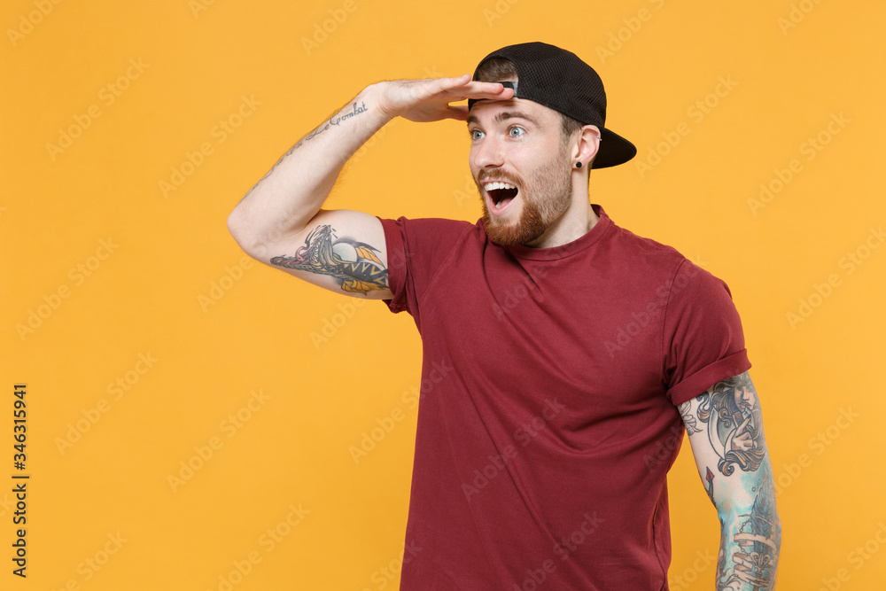 Excited young bearded tattooed man guy in casual t-shirt, black cap posing isolated on yellow background. People lifestyle concept. Mock up copy space. Hold hand at forehead looking far away distance. Excited young bearded tattooed man guy in casual t-shirt, black cap posing isolated on yellow background. People lifestyle concept. Mock up copy space. Hold hand at forehead looking far away distance.