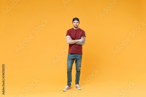 Attractive young tattooed man guy in casual t-shirt black cap posing isolated on yellow wall background studio portrait. People emotions lifestyle concept. Mock up copy space. Holding hands crossed.