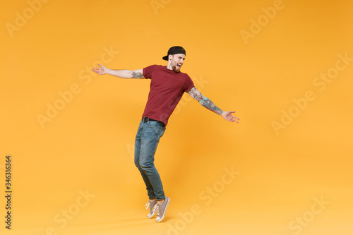 Young tattooed man guy in casual t-shirt black cap posing isolated on yellow wall background studio portrait. People lifestyle concept. Mock up copy space. Dancing  standing on toes  spreading hands.