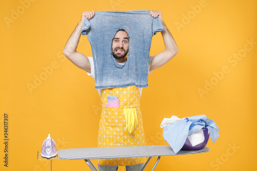 Confused young man househusband in apron ironing clean clothes on board while doing housework isolated on yellow wall background studio. Housekeeping concept. Mock up copy space. Hold burnt t-shirt. photo