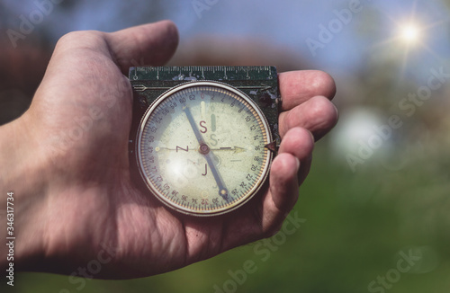 Man holding an old compass in one hand. Male holding a military compass showing direction. Blue sky and sun in the background, first person point of view, close up.