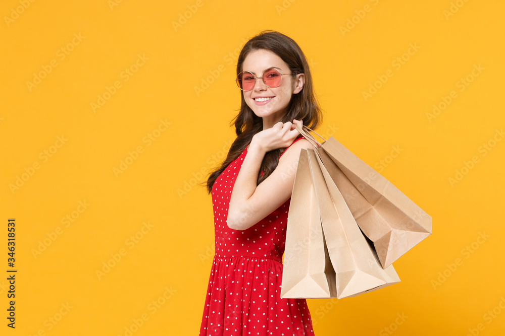Smiling young brunette woman girl in red summer dress, eyeglasses posing isolated on yellow wall background studio portrait. People lifestyle concept. Hold package bag with purchases after shopping.