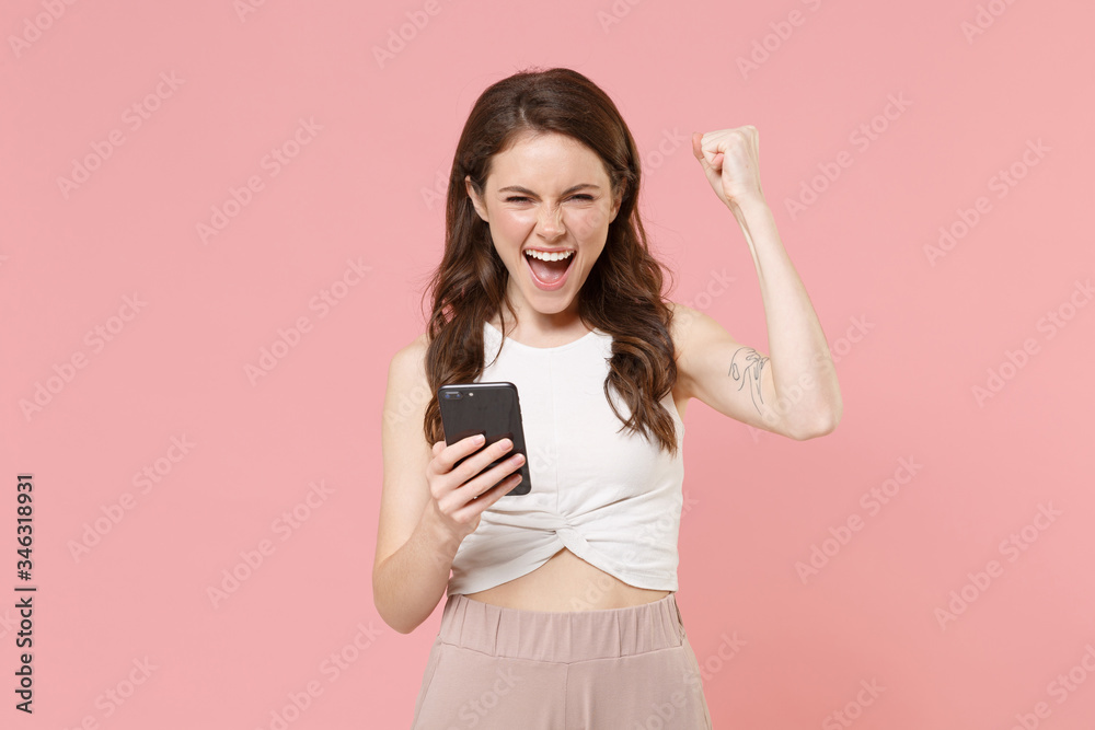 Happy young brunette woman girl in light casual clothes isolated on pastel pink background. People lifestyle concept. Mock up copy space. Using mobile phone, typing sms message, doing winner gesture. Happy young brunette woman girl in light casual clothes isolated on pastel pink background. People lifestyle concept. Mock up copy space. Using mobile phone, typing sms message, doing winner gesture.