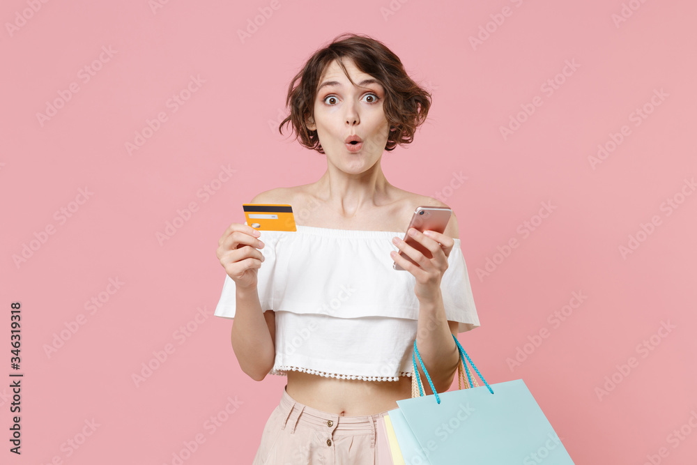 Fototapeta premium Shocked young woman girl in summer clothes isolated on pink wall background. Shopping discount sale concept. Mock up copy space. Using mobile phone, hold package bag with purchases, credit bank card.