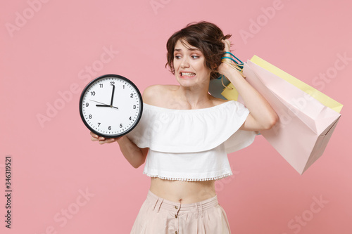 Confused puzzled young woman girl in summer clothes isolated on pastel pink wall background studio portrait. Shopping discount sale concept. Mock up copy space. Hold package bag with purchases, clock.