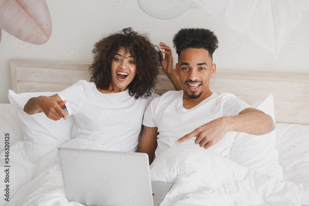 Young calm african loving couple girl guy in white t-shirts using laptop pc computer lying on bed indoors in bedroom at home, spending time in room. Rest relax good mood quarantine lifestyle concept.