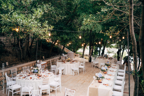 Fototapeta Naklejka Na Ścianę i Meble -  Wedding dinner table reception. Round tables with white tablecloths, brown runners, bouquets flowers, Chiavari chairs, menu sin-enclosed in napkin. Restaurant outside, in woods, water lights garlands
