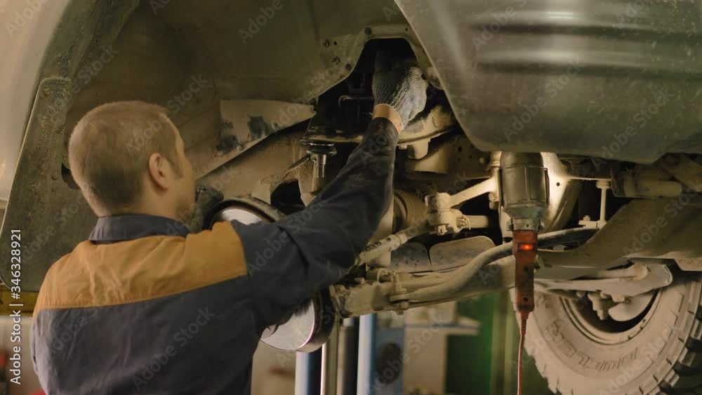 An Asian man is repairing the chassis of an old car lifted on a lift in a workshop, close-up. An Asian man is repairing the chassis of an old car lifted on a lift in a workshop, close-up.