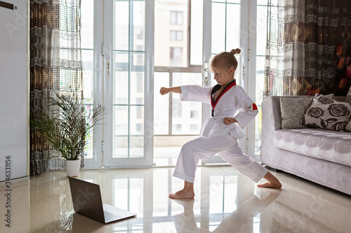 Taekwondo girl in kimono with white belt exercising at home in living room. Online education during coronavirus covid-19 lockdown, self isolation and social distancing concept.