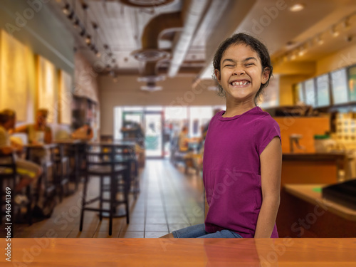 A funny young girl gives the camera a super excited smile inside the coffeehouse. Casually dressed and fully erected on a stool with people enjoying their coffee in the background.