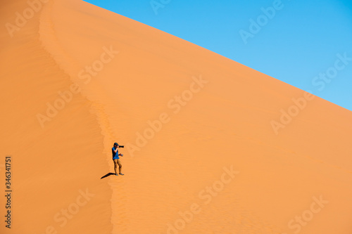 Asian man photographer hold a camera walking to sand Dune in hot temperature at noon with clear blue sky. A popular landmark for tourist in NAMIBIA.