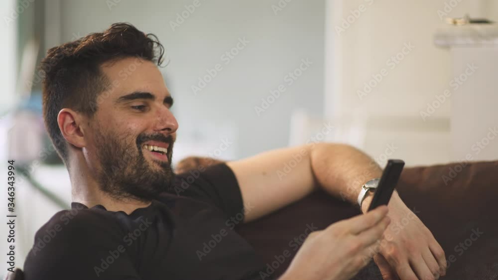 Caucasian man smiling, black shirt chilling at home on brown sofa inside, during covid-19 pandemic in France. 4K UHD. One person young man having phone conversation with smartphone. Caucasian man smiling, black shirt chilling at home on brown sofa inside, during covid-19 pandemic in France. 4K UHD. One person young man having phone conversation with smartphone.