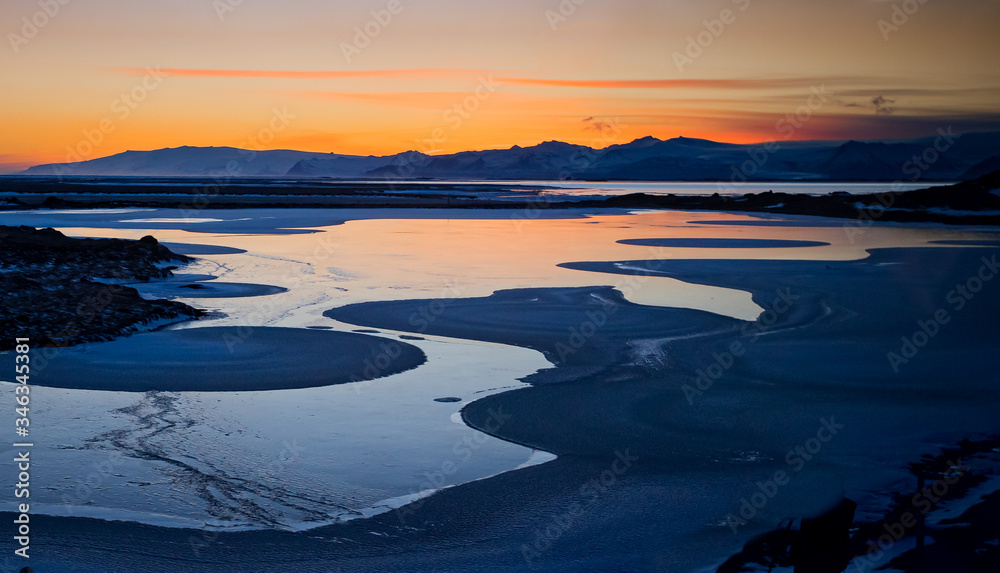 Dramatic sunset reflects orange color into tidal pools near black beach and Vestrahorn