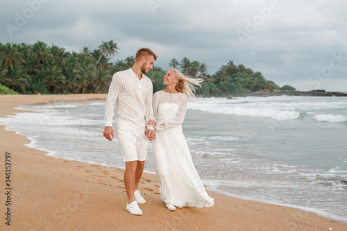 Happy groom and beautiful bride  walking  on tropical sand beach wih palms. Sea on the background.... © Yevhenii