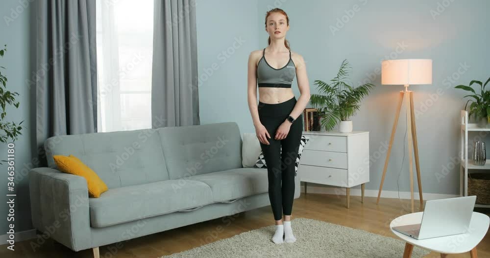 Young Caucasian sporty girl working out for legs in cozy living room during lockdown. Beautiful red-haired female exercising for muscles while lifting leg at home. Self-isolation concept. Young Caucasian sporty girl working out for legs in cozy living room during lockdown. Beautiful red-haired female exercising for muscles while lifting leg at home. Self-isolation concept.