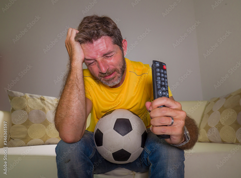 Obraz premium frustrated football fan in intense emotion - home portrait of young dejected and sad man watching soccer game on television at living room couch his team losing