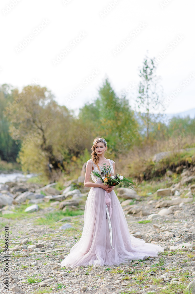 Happy bride in a pink wedding dress. The girl holds a wedding bouquet in her hands. Boho style wedding ceremony near the river.