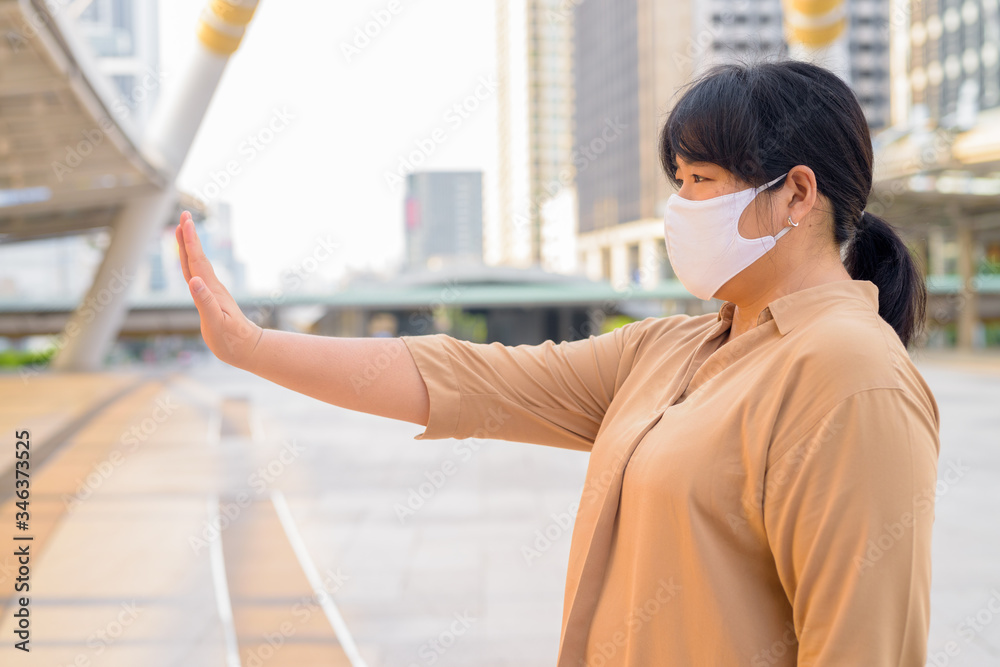Profile view of overweight Asian woman with mask showing stop gesture in the city Profile view of overweight Asian woman with mask showing stop gesture in the city