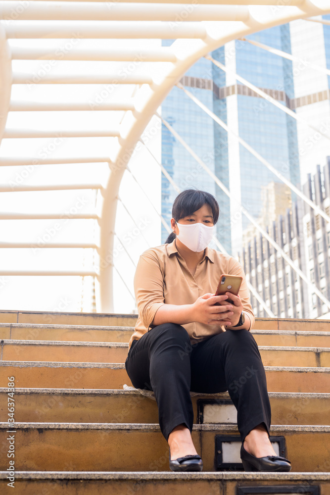 Overweight Asian woman with mask using phone and sitting in the city Overweight Asian woman with mask using phone and sitting in the city