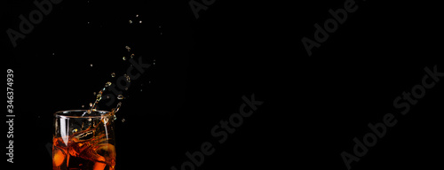 An ice cube falls into a glass with whiskey and the spray flies beautifully in different directions on a black background. Panoramic photo for a banner