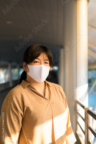 Overweight Asian woman with mask thinking at footbridge in the city