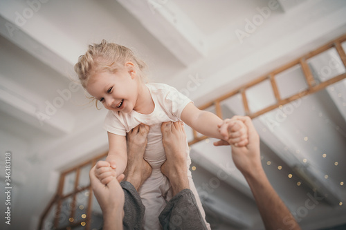 very beautiful blonde preschool girl is playing with an adult in a cozy white room with a high ceiling. men's legs hold the baby up like a plane. activities with children at home