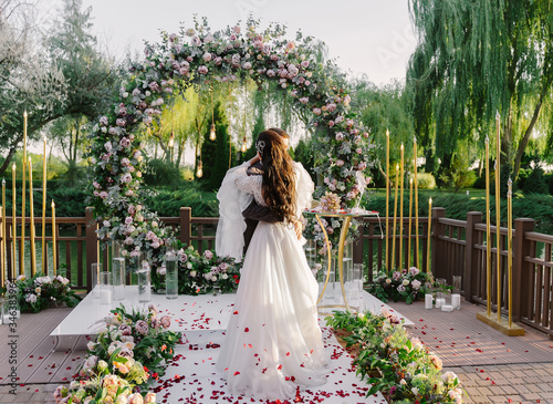 Newlywed bride and groom hugging and kissing at the wedding ceremony on the background of round wedding arch decorated with roses and eucalyptus leaves, guests are tossing red roses petals at them.