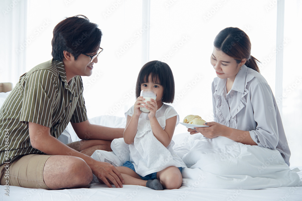 Happy Asian family having breakfast with milk bread sausages at bedroom, a little daughter drinking a glass of milk on bed at home Happy Asian family having breakfast with milk bread sausages at bedroom, a little daughter drinking a glass of milk on bed at home
