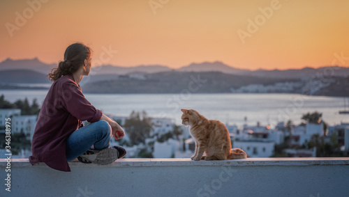 A young girl sits on a roof at sunset and looks at the evening city and the sea with her red cat