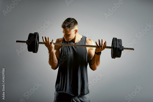 A man does exercises with a barbell on a gray background. The athletic body of a young man in muscle tension makes an approach with a barbell. Copy space