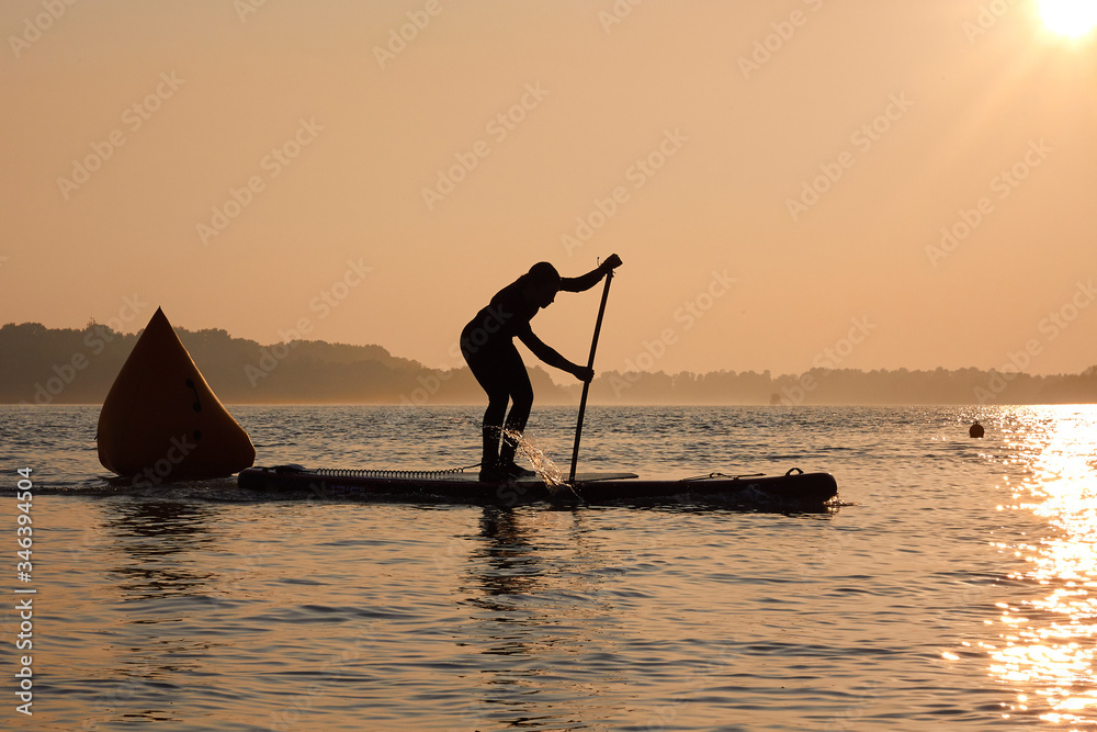 SUP silhouette of teenager standing with a paddle on the surfboard at sunset. Stand up paddle boarding competitions on the calm river