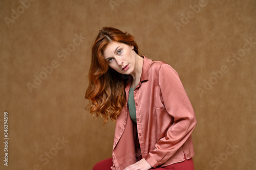A pretty model with long hair is dressed in red trousers and a pink blouse. Portrait of a young Caucasian red-haired woman sitting on a stand on a beige background in various poses.