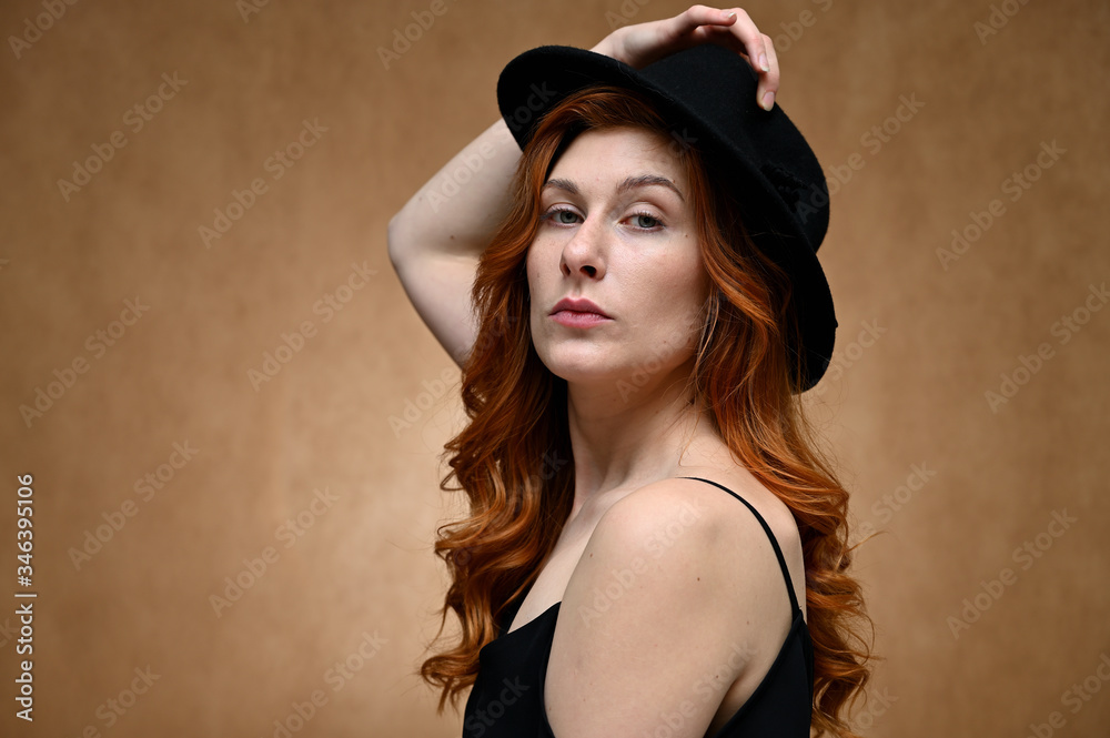 Close-up portrait of a young pretty woman with long red hair on a beige background. Model posing in a black hat. Close-up portrait of a young pretty woman with long red hair on a beige background. Model posing in a black hat.