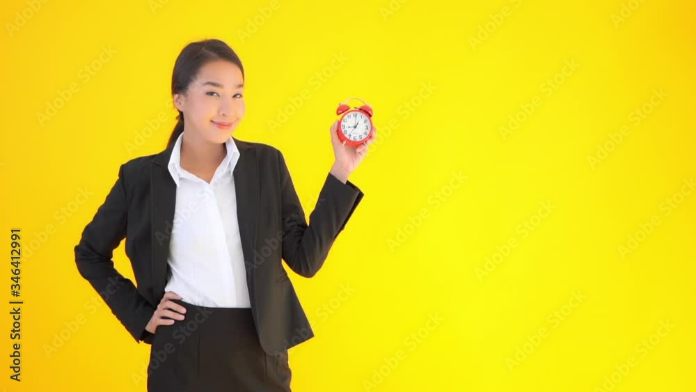 Attractive Asian woman in business attire holding red alarm clock with bells points to time. Tardy, late for work, business deadline concept. Bright yellow background and lady with clock. Attractive Asian woman in business attire holding red alarm clock with bells points to time. Tardy, late for work, business deadline concept. Bright yellow background and lady with clock.