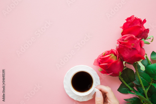 Young woman hand holding white cup of black coffee. Red artificial roses. Empty place for inspirational, sentimental text, lovely quote or sayings on light pink table background. Pastel color. Closeup