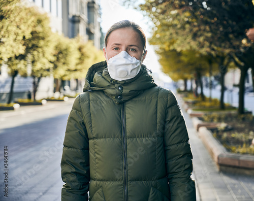 Portrait of a woman in a mask, outside in an empty street, green trees in sunlight on a background. Social distancing during global coronavirus quarantine