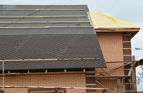 A close-up on a roofing construction with asphalt roof shingles installation on the waterproof underlayment and uncovered roof sheathing of an incomplete house construction with scaffoldings.