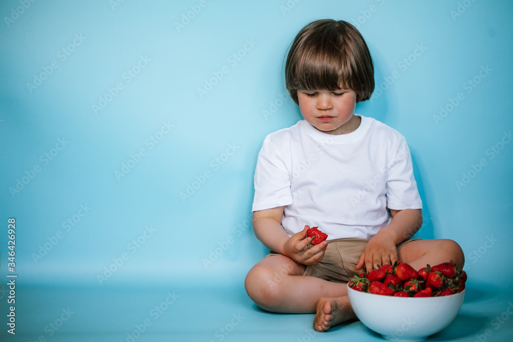 Toddler boy sitting with bowl of strawberries on the blue background Toddler boy sitting with bowl of strawberries on the blue background