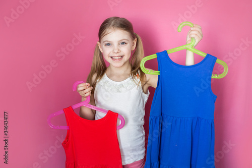 Baby on a pink background, little girl holds two dresses of different colors, rejoices in buying two for the price of one