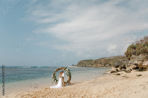 Romantick wedding cople standing on the beach near round wooden arch in boho rustc style  after ceremony by the sea. Moment of tenderness and love. Beach wedding Bali photo