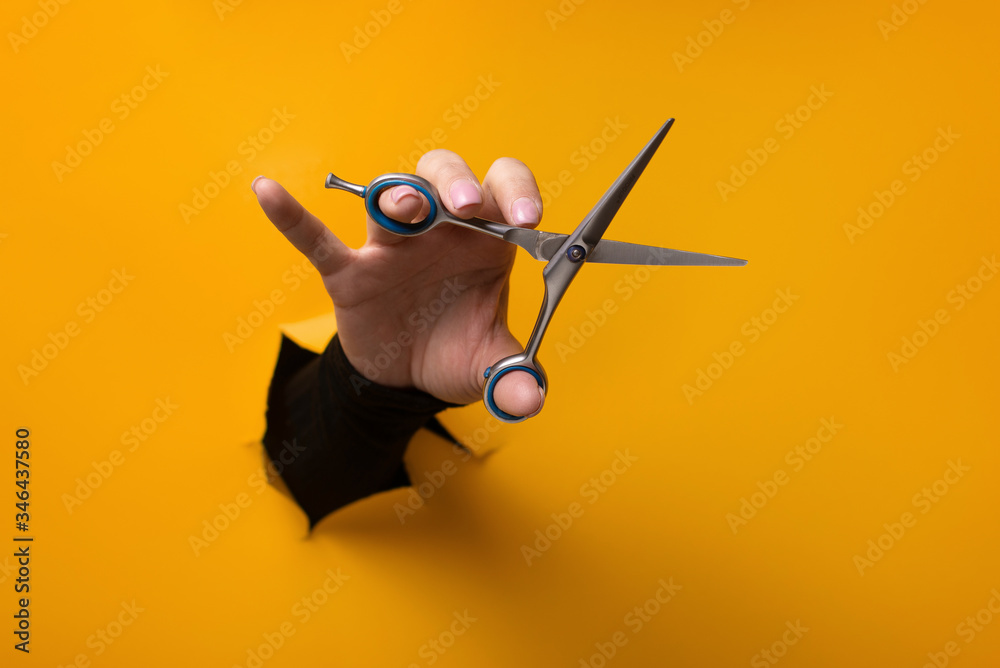 hairdresser holding in hand scissors for cutting hair from a torn hole in orange paper - obrazy, fototapety, plakaty 