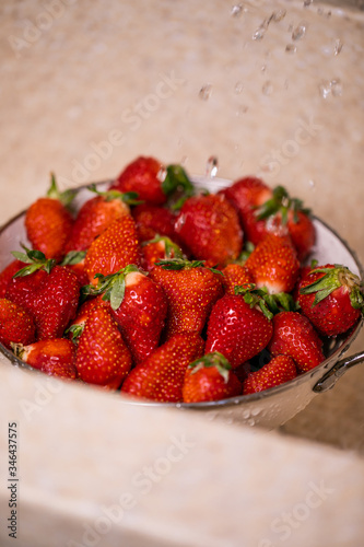 Ripe strawberries in white colander standing under pouring water in sink