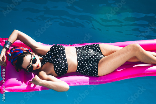 
Beautiful girl by the pool with long dark hair and make-up resting at a spa resort. In a black retro bathing suit, scarf, hat and glasses, smiles sunbathes on a pink 
inflatable mattress in the motel