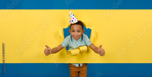 Сheerful and happy birthday boy African-American with cone cap on his head against ragged paper yellow background of wall, reaching out to hold something or take it, asks to pick it up.