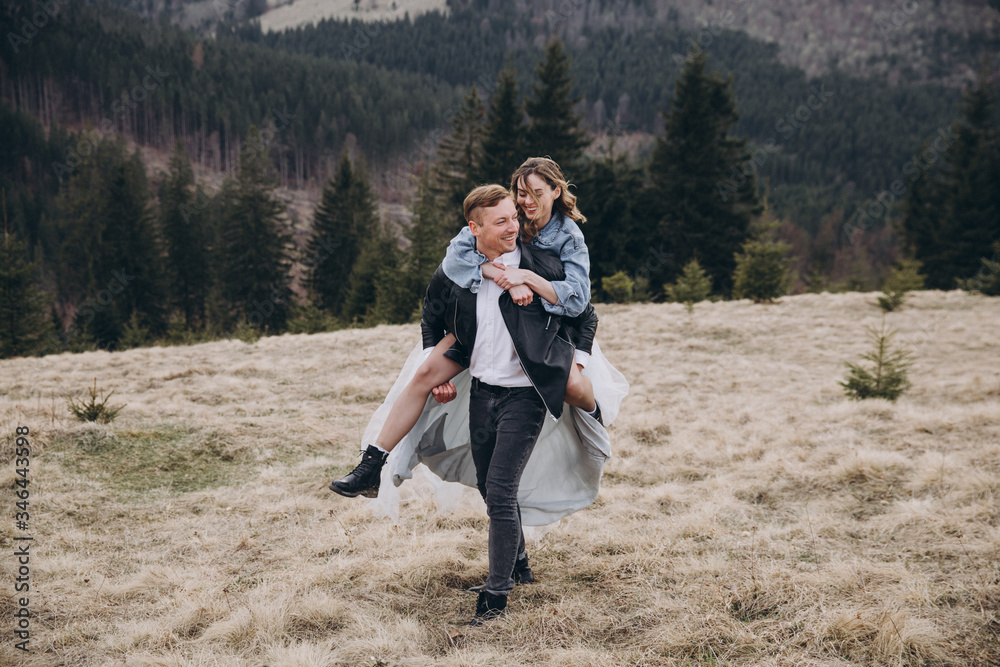 Stylish couple in the autumn mountains. A guy in a leather jacket and a young girl in a gray blue wedding dress and a denim jacket having fun together on the background of a sky, forest and landscapes